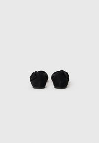 Black suede slip-on shoes featuring a rounded toe and bow accent on the side. Smooth texture with minimal heel height.