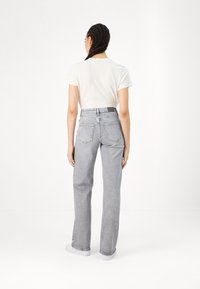 Femme aux cheveux tressés portant une chemise blanche à manches courtes rentrée dans un jean gris taille haute, jambes droites, vue de dos sur fond blanc.