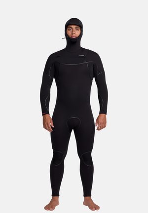 Homme debout pieds nus, portant une combinaison de plongée noire intégrale avec capuche, face à un fond clair et uni.