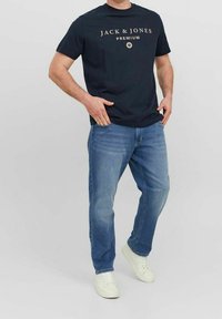 Camiseta azul marino con texto de logo dorado; vaqueros de mezclilla azules; zapatillas blancas; ajuste casual con mangas cortas y diseño de pierna recta.