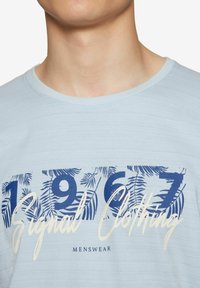 Šviesiai mėlynas medvilninis marškinėlis su apvaliu iškirpimu, ant kurio yra spausdinta dizaino su tamsiai mėlynomis skaičiais "1967" ir "Signal Clothing" baltais kursyviniais rašmenimis.
