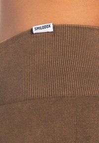 Zbliżenie brązowej materiału prążkowanego z białą metką "SMILODOX" wszytą w pasie, na tle gładkiej skóry.