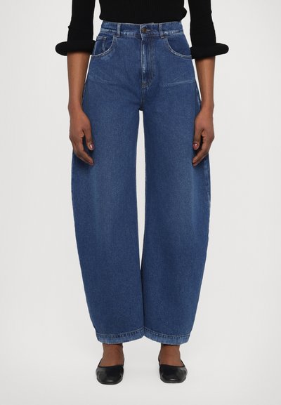 Jeans a vita alta e gambe larghe in denim blu medium. Presentano tasche frontali, chiusura a zip e orlo pulito. Indossati con ballerine nere.