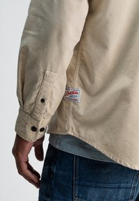 Beige corduroy overhemd met een knoopmanchet, voorzien van een rood-wit merklabel op de zijnaad. Draag het over een grijs T-shirt en blauwe jeans.