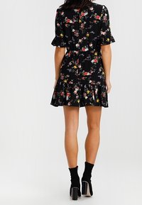 Kvinna i en svart blommig kort klänning med volangkant och ärmar, iklädd svarta klackade ankelskor, stående med ryggen vänd.