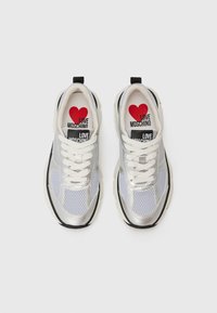 Srebrni in beli športni copati z mrežasto zgornjim delom, teksturiranimi črnimi detajli, belimi vezalkami in rdečim srcem na podplatu z napisom "LOVE MOSCHINO."