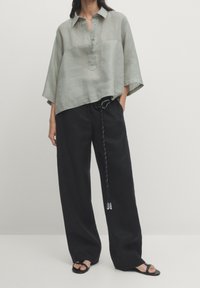 Loose-fitting, short-sleeve, grey linen top with a front button placket, paired with high-waisted black wide-leg trousers and black sandals.