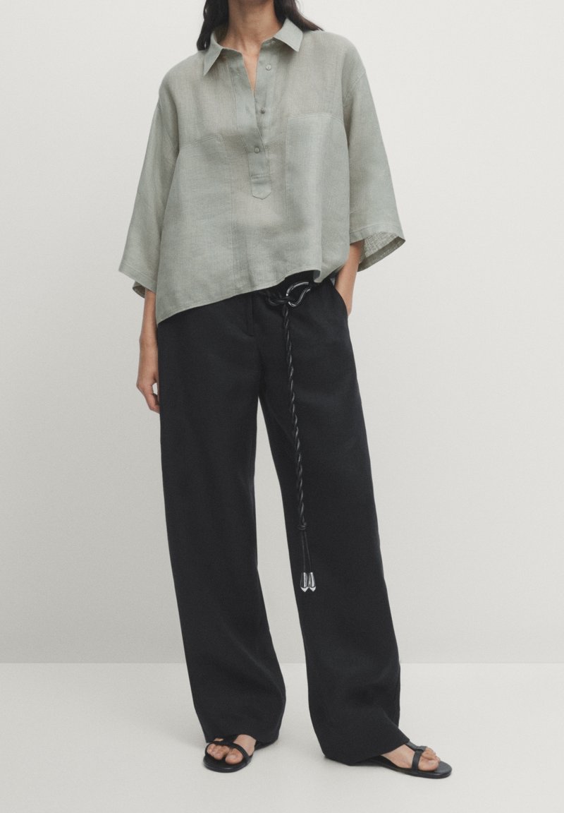 Loose-fitting, short-sleeve, grey linen top with a front button placket, paired with high-waisted black wide-leg trousers and black sandals.