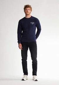 Felpa blu navy con logo bianco "PTRL", girocollo e polsini a coste, abbinata a jeans neri e sneakers bianche. Design semplice, vestibilità casual.