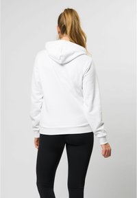 Hvit hettegenser med myk, strukturert tekstur og ribbede mansjetter. Brukt med sorte leggings, som viser en avslappet bakvinkel.