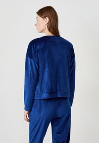 Kobieta z brązowymi włosami, ubrana w luźną, długą, welurową bluzkę w kolorze granatowym oraz pasujące spodnie, widoczna z tyłu na białym tle.