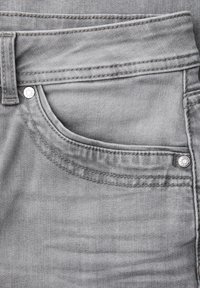 Gros plan sur une poche de jean en denim gris clair avec double couture, rivets métalliques et passants de ceinture sur le côté supérieur gauche.