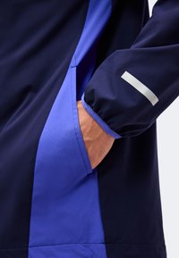 Main dans la poche de la manche d'une veste bleu marine et bleue avec une bande réfléchissante sur le poignet contre un fond uni.