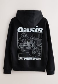 Zwarte sweatshirt met capuchon, voorzien van een wit lijntekeninggrafiek op de achterkant, met de tekst "oasis" en "BE HERE NOW" duidelijk zichtbaar.