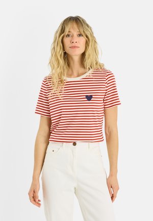 Mujer con cabello rubio ondulado que lleva una camiseta de manga corta a rayas rojas y blancas con un pequeño corazón azul marino y pantalones blancos de talle alto.
