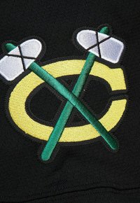 Lettre "C" jaune avec deux crosses de hockey vertes croisées et des lames enroulées de blanc sur fond de tissu noir.