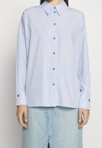 Camicia a maniche lunghe a righe blu e bianche con colletto, bottone frontale e maniche con polsini, abbinata a una gonna di jeans.