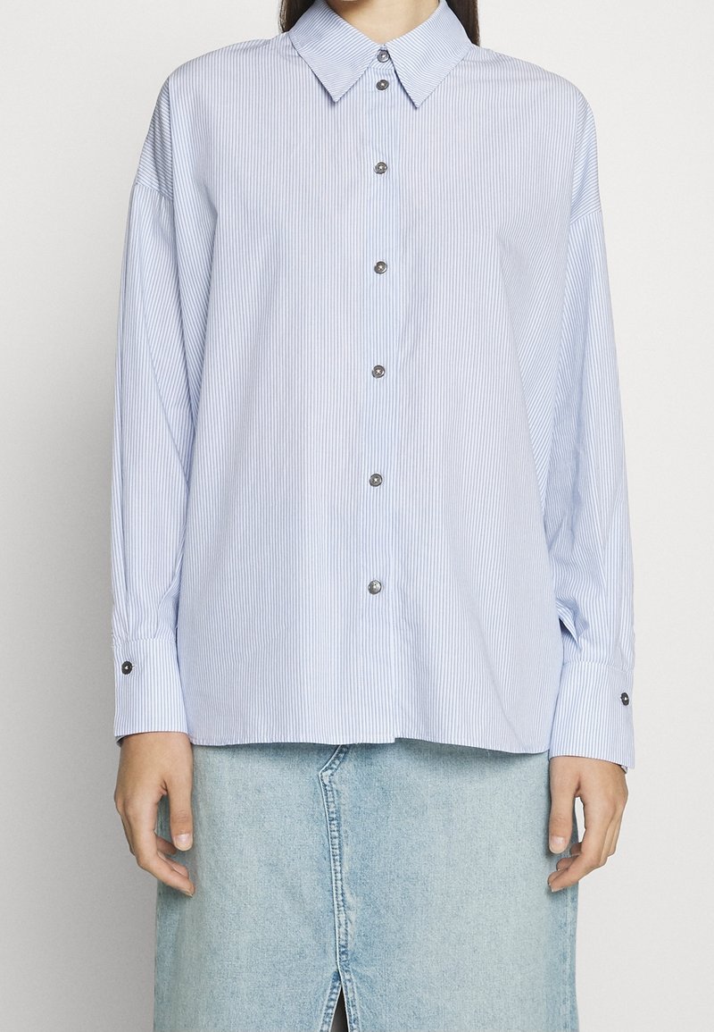 Camicia a maniche lunghe a righe blu e bianche con colletto, bottone frontale e maniche con polsini, abbinata a una gonna di jeans.