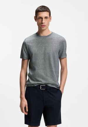 Homme debout, les mains dans les poches, portant un t-shirt gris à manches courtes et un short bleu marine avec une ceinture noire, sur fond blanc.