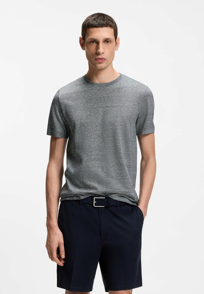 Mann steht mit den Händen in den Taschen, trägt ein graues kurzärmeliges T-Shirt und marineblaue Shorts mit schwarzem Gürtel, vor weißem Hintergrund.