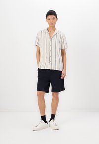 Camicia a maniche corte con bottoni a righe crema e blu navy, abbinata a pantaloni corti scuri. Modello che indossa sneaker bianche e calze nere.