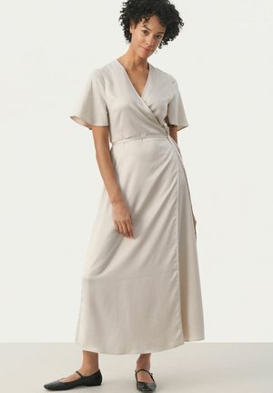 Beige wrap dress in smooth fabric, featuring short sleeves, a belted waist, and a side slit, paired with black flat shoes.
