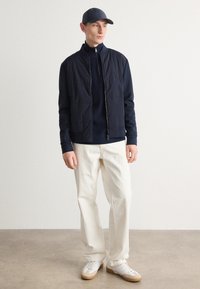 Navy zip-up jacket over a navy sweater, paired with cream trousers and white sneakers with tan accents; set against a neutral background.