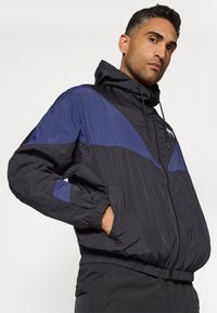 Veste coupe-vent noire en nylon avec un design en blocs de couleurs diagonales bleues, dotée d'une capuche, de poignets élastiques et de poches frontales zippées.