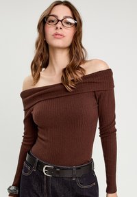 Brun ribbet off-the-shoulder top med lange ærmer, der har et tætsiddende design og struktureret stof. Kombineret med et sort bælte og mørk denim.