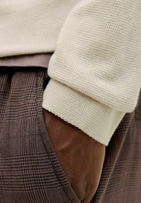 Main à la peau foncée dans la poche d'un pantalon à carreaux marron, partiellement couverte par une manche de pull en tricot couleur crème.