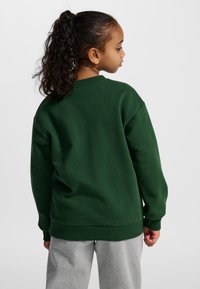 Grön sweatshirt av tjockt tyg med ribbstickade muddar och fåll, bärs med gråa sweatpants. Designen har sänkta axelsömmar.