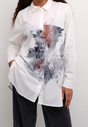 Femme portant une chemise blanche à boutons avec des imprimés floraux abstraits noirs et rouges, associée à un pantalon noir, mains dans les poches.