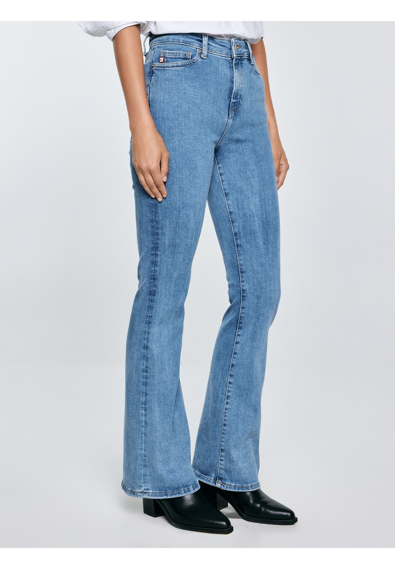 Žena oblečená v světle modrých mírně rozšířených džínách a černých kotníkových botách na podpatku, stojící na jednoduchém světlém pozadí.