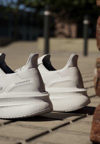 Coppia di sneakers bianche Adidas Ultraboost posizionate sul marciapiede all'aperto al sole, con attenzione ai dettagli del tallone e dei lati.