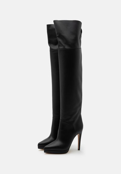 Bottes hautes en cuir noir avec bouts pointus et talons aiguilles fins, debout droit sur un fond uni.