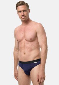 Mężczyzna bez koszulki, o krótkich blond włosach, ubrany w granatowe slipy z czarnym pasem, stojący na tle jasnego, jednolitego tła.