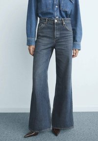 Persona vistiendo jeans de campana oscuros de cintura alta, combinados con una camisa de denim azul medio metida por dentro y zapatos marrones de punta.