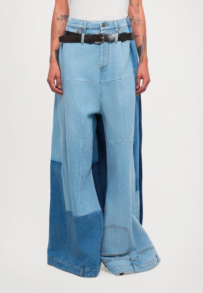 Pantalon en denim bleu clair à jambes larges avec un design en patchwork, des bords bruts et une ceinture noire. Le tissu présente des nuances de bleu variées.