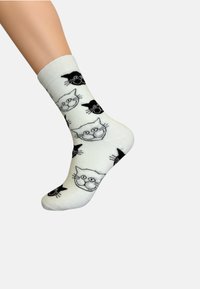 Vita bomullssockor med ett upprepande mönster av svarta katthuvuden med morrhår. Designen innehåller lekfulla ansiktsuttryck.