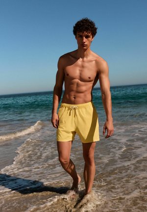 Junger Mann mit lockigem Haar, der gelbe Badeshorts trägt und barfuß auf nassem Sand am Meer entlang läuft, unter klarem blauem Himmel.