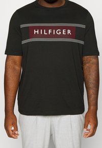 Musta lyhythihainen t-paita, jossa on punainen ja valkoinen "HILFIGER" -logografiikka keskellä, ohuiden raitojen kanssa ylhäällä ja alhaalla. Pehmeä kangas.