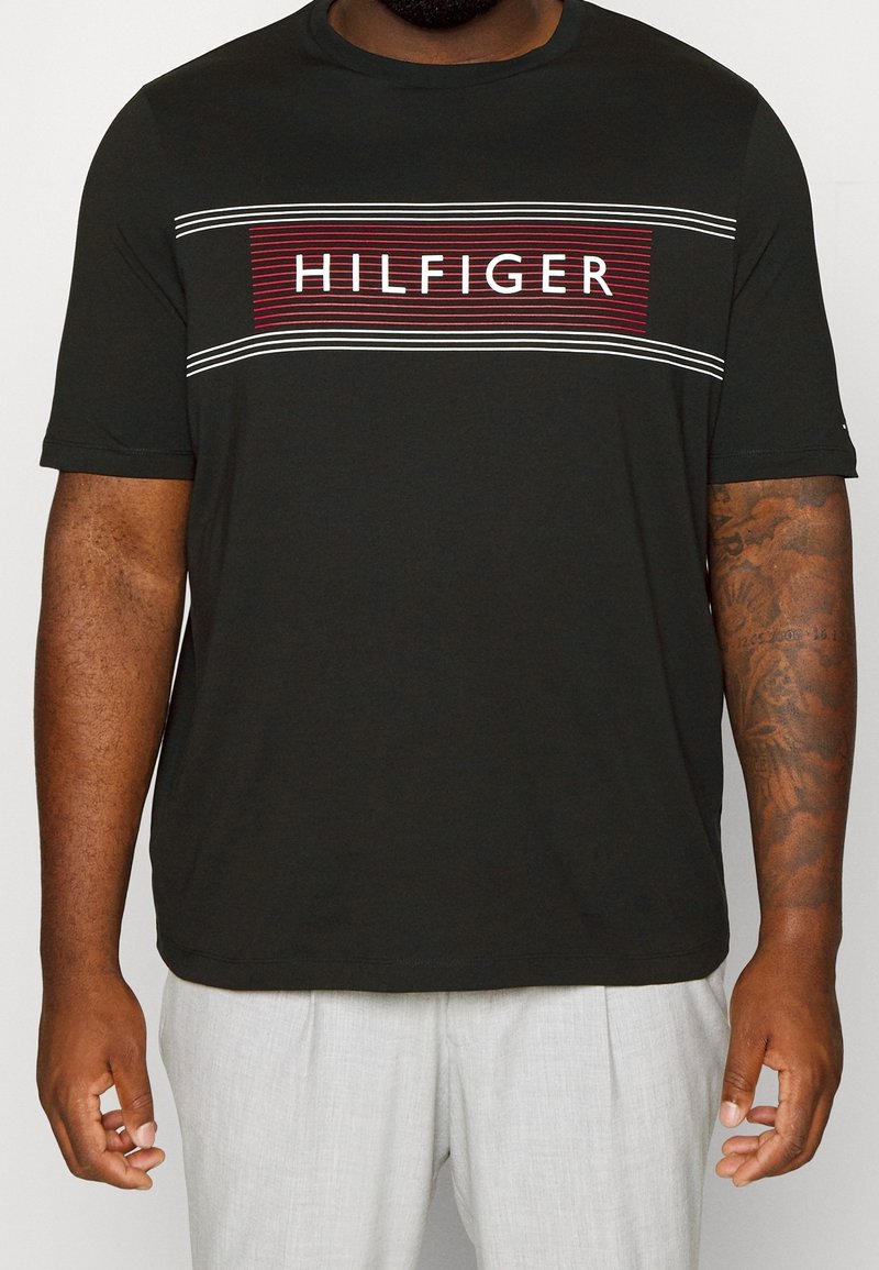 Musta lyhythihainen t-paita, jossa on punainen ja valkoinen "HILFIGER" -logografiikka keskellä, ohuiden raitojen kanssa ylhäällä ja alhaalla. Pehmeä kangas.