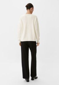 Weißer, gerippter Strickpullover mit rundem Halsausschnitt und vertikaler Nahtdetail auf dem Rücken, kombiniert mit schwarzen, weit geschnittenen Hosen und Stiefeletten.