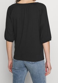 En person som bär en svart off-shoulder-blus med trekvartsärmar och ljusblå denimjeans, sedd bakifrån.