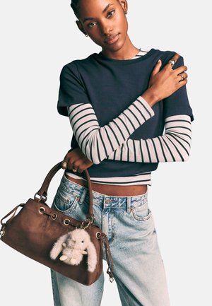 Borsa in suede marrone con un portachiavi a forma di coniglietto bianco e peloso; la modella indossa una maglietta cropped blu navy sopra una maglietta a maniche lunghe a righe e jeans chiari.