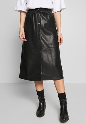 Personne portant une jupe midi en cuir noire à boutons sur le devant, une chemise blanche, des chaussettes noires et des bottines noires brillantes, debout sur un sol gris.