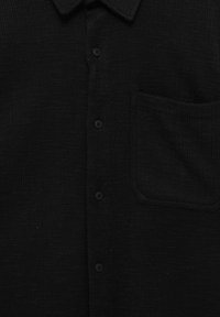 Camisa negra de tejido trenzado con un tejido texturizado, cierre de botones y un bolsillo en el pecho. El cuello es estructurado y plano.