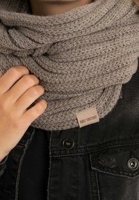 Gehaakte grijze sjaal met een dik, gestructureerd patroon. Een leren label met "KNIT FACTORY" is bevestigd en wordt gedragen over een zwarte spijkerjack.