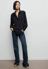 Svart skjorta med knappar, långa ärmar, avslappnad passform; kombinerad med utsvängda mörka jeans. Jeansen har en jämn textur och minimal detaljer.