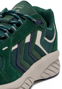 Grön sportig sko med mocka och mesh-material, med en texturerad grå sula. Anmärkningsvärd design inkluderar zigzag-detaljer och vadderad krage.